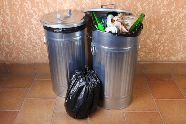 Two aluminum garbage cans full of garbage, including bottles and cloth. Black plastic garbage bag sitting between the two cans.